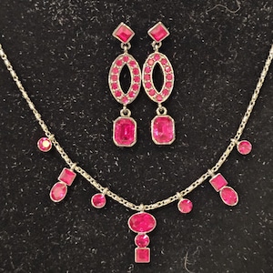 May include: A silver necklace and matching earrings set. The necklace features a series of pink gemstones in various shapes, including circles and squares, suspended from a delicate chain. The earrings have oval and rectangular pink gemstones.