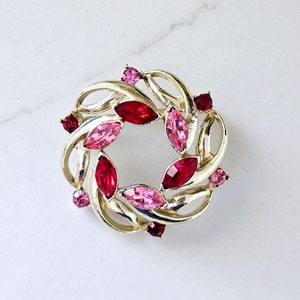 May include: A vintage gold-tone brooch with a circular design. The brooch features a wreath-like pattern with alternating pink and red marquise-cut rhinestones. Small pink rhinestones are also scattered throughout the design.