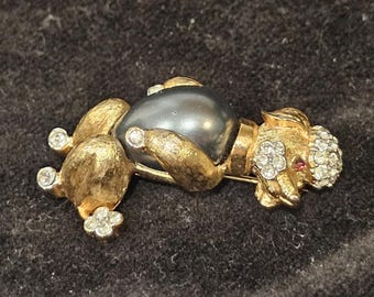 May include: A gold-coloured poodle brooch with a dark grey pearl body. The poodle's head and legs are decorated with clear rhinestones and a small red accent. A vintage jewellery item.