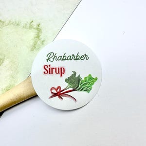 Könnte beinhalten: Ein rundes, weißes Etikett mit den Worten "Rhabarber Sirup" in Grün und Rot. Das Etikett zeigt eine Aquarellillustration einer Rhabarberpflanze mit grünen Blättern und einer roten Schleife. Das Etikett befindet sich auf einer weißen Oberfläche.