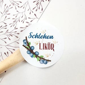 Könnte beinhalten: Ein weißer, runder Aufkleber mit den Worten "Schlehen LIKÖR" in Blau und Rot, mit goldenen Akzenten. Auf dem Aufkleber ist eine Aquarellillustration eines Zweigs mit blauen Beeren abgebildet. Der Hintergrund umfasst gemustertes Papier und einen Holzrundstab.