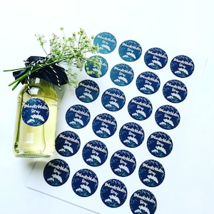 Könnte beinhalten: Eine Glasflasche mit klarer Flüssigkeit, verziert mit einem marineblauen Etikett mit weißen Blumenillustrationen und dem Text "Holunderblüten Sirup". Mehrere passende runde Aufkleber sind in der Nähe verstreut. Die Flasche ist mit schwarzem Band und Grünzeug gebunden.