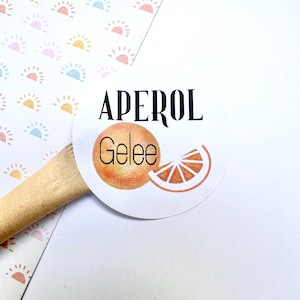 Könnte beinhalten: Ein runder weißer Aufkleber mit dem Text "APEROL Gelee" und einer Orangenscheiben-Illustration. Der Aufkleber befindet sich auf einer weißen Oberfläche mit einem Holzlöffel und gemustertem Papier mit Sonnenmotiven.