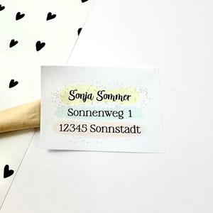 Könnte beinhalten: Ein weißes rechteckiges Etikett mit dem Namen "Sonja Sommer" in schwarzer Schrift, der Adresse "Sonnenweg 1" und der Postleitzahl "12345 Sonnstadt". Das Etikett hat pastellgelbe, blaue und rosa Akzente. Schwarze Herzformen sind im Hintergrund verstreut.