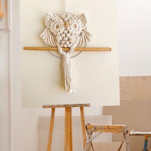 May include: A white macrame owl wall hanging with a wooden dowel and a tassel. The owl is hanging on a white wall with a wooden easel in the foreground.