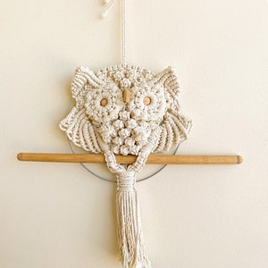 Könnte beinhalten: Ein Makramee-Wandbehang in Form einer Eule mit einem Holzstab und einem Quasten. Die Eule ist aus naturfarbenem Garn gefertigt und hat Holzaugen.