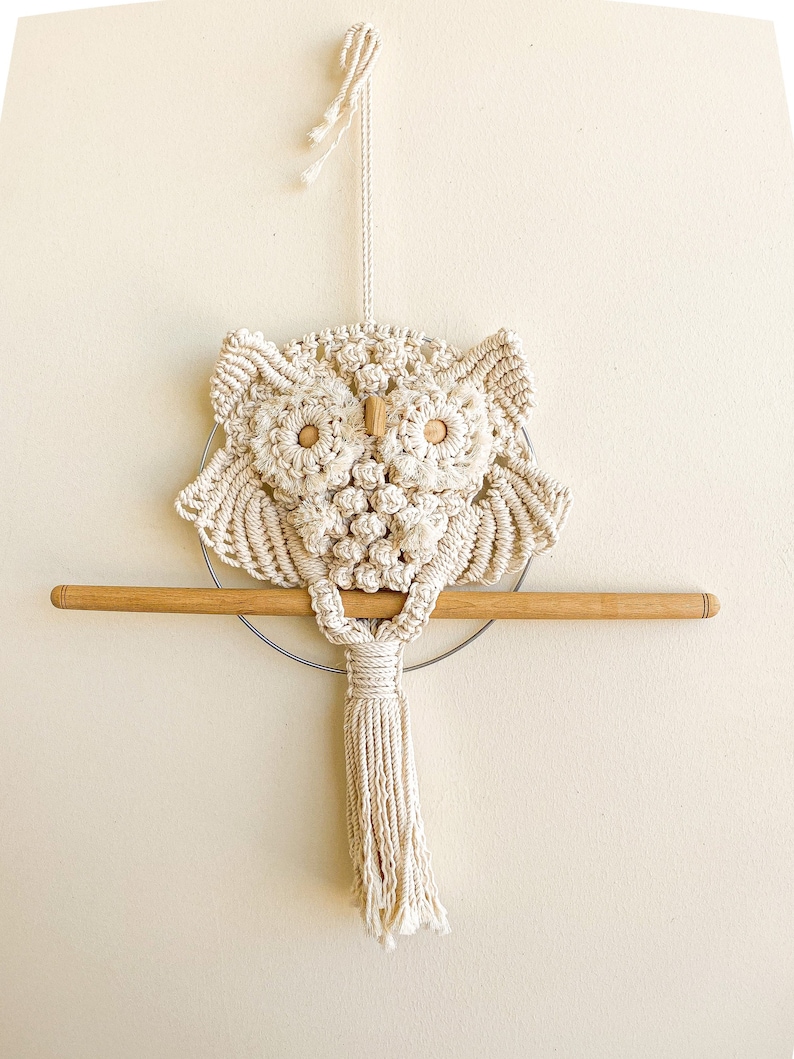 May include: A macrame owl wall hanging with a wooden dowel and a tassel. The owl is made with natural coloured string and has wooden eyes.