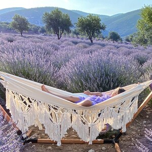 May include: A white hammock with fringe hanging from the bottom is strung between two wooden posts. A person is lying in the hammock, which is set in a field of lavender.