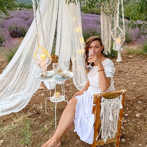 May include: A woman in a white dress sits at a table with a white tablecloth and a white metal stand. The table is set with two plates, two glasses, and a small bowl. The woman is holding a glass of wine and looking at the camera. The table is decorated with a white macrame hanging and a white macrame chair back. The scene is set in a field of lavender.
