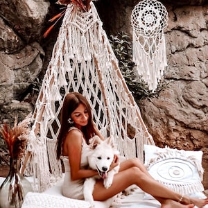 Op de afbeelding: Een vrouw zit in een witte macramé tipi met een witte hond. Ze draagt een witte jurk en houdt de hond vast. Er hangt een witte droomvanger aan de tipi.