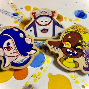 May include: Three wooden character pins. One pin features a character with blue hair and red eyes. Another pin features a character with a white face and a purple hat. The third pin features a character with yellow hair and brown skin.