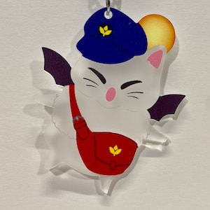 May include: A white cartoon cat-like character with a blue hat, purple wings, and a red satchel. The character has a pink nose and is smiling. A yellow sun is behind the character. The keychain is attached to a silver chain.