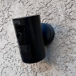 Może przedstawiać: Czarna kamera bezpieczeństwa Ring zamontowana na białej ścianie z tynku. Kamera ma czarny obiektyw i białe logo "ring".