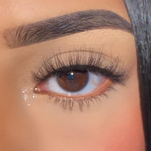 Op de afbeelding: Close-up van een bruin oog met lange, donkere wimpers en een goed gedefinieerde wenkbrauw. Het oog reflecteert licht en er zijn kleine, glinsterende druppels in de buurt van de onderste wimperlijn. De huid rond het oog heeft een zachte, perzikkleurige tint.