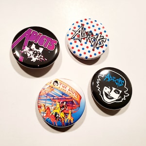 May include: Four round pinback buttons with different designs. One button has the word "ADICTS" in purple, another has the word "THE ADICTS" in a dotted pattern, and the other two have illustrations of a carnival ride and a smiling face.