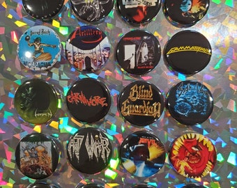 Op de afbeelding: Een verzameling van twintig zwarte buttonspins met metalbandlogo's en albumhoezen. De ronde pins tonen ontwerpen in verschillende kleuren, waaronder rood, blauw en wit. De pins hebben een glanzende afwerking.