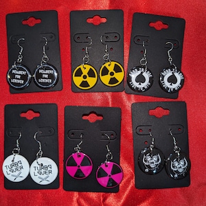 Puede incluir: Seis pares de pendientes con diferentes diseños. Los pendientes se muestran en cartulina negra. Los diseños incluyen una calavera en blanco y negro, una pica en blanco y negro con el texto "Born to Lose, Live to Win", una calavera en blanco y negro con el texto "Turbo Lover", un símbolo de radiación en blanco y negro, un símbolo de radiación amarillo y negro, y un círculo en blanco y negro con el texto "Wellbent for Leather".