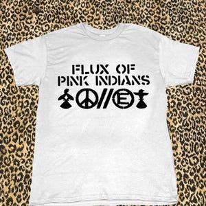Könnte beinhalten: Ein weißes T-Shirt mit dem schwarzen Text "FLUX OF PINK INDIANS". Darunter befinden sich drei schwarze Symbole: eine Person, ein Friedenszeichen und ein Symbol mit dem Buchstaben "E". Das T-Shirt wird vor einem Leopardenmuster-Hintergrund gezeigt.