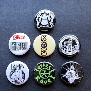 Könnte beinhalten: Neun schwarze und weiße Anstecknadeln mit verschiedenen Designs. Die Nadeln zeigen Bandnamen und Logos, darunter "RATTUS", "DESTRUCKTIONS", "KAAOO", "Terveet Kädet", "LAMA" und "MOB 47".