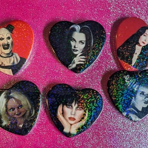 May include: Six heart-shaped pins featuring horror movie characters. The pins have a glittery background and include images of a clown, Morticia Addams, Elvira, Chucky, and the Bride of Frankenstein.