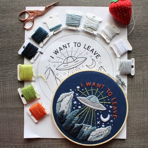 Peut inclure: Un cerceau de broderie blanc avec un fond bleu et un motif brodé coloré d'un OVNI volant au-dessus des montagnes avec le texte "I want to leave". Le cerceau est entouré de fils à broder de différentes couleurs.