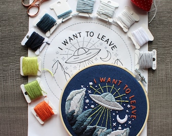 May include: A white embroidery hoop with a blue background and a colorful embroidered design of a UFO flying over mountains with the text "I want to leave". The hoop is surrounded by various colors of embroidery floss.