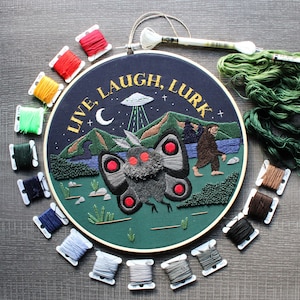 Puede incluir: Arte de bordado en aro con un fondo verde oscuro que presenta una polilla con ojos rojos y el texto "Live, Laugh, Lurk". El aro está rodeado de carretes de hilo de bordar en varios colores.