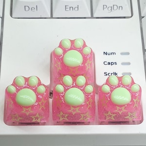 May include: Three custom mechanical keyboard keycaps shaped like cat paws. Each keycap features a translucent pink base with embedded gold star shapes and a raised, smooth, light green paw pad and toe beans on the top surface.