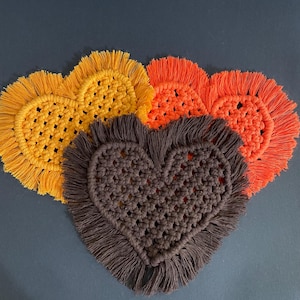 May include: Three heart-shaped macrame coasters featuring a knotted cord pattern and a fringed edge. The coasters are colored in mustard yellow, bright orange, and dark brown, arranged in a layered cluster against a dark gray background.