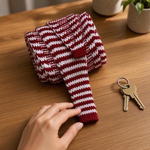 Peut inclure: Une cravate en tricot enroulée présentant un motif répétitif de rayures horizontales rouge foncé et blanches. Le tissu a un aspect texturé et côtelé. Un petit porte-clés en métal avec deux clés dorées est posé à proximité.