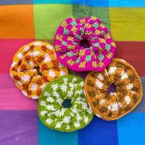 May include: Four handmade crochet scrunchies in various colors. One is bright pink with green and yellow accents, another is green with white, and the others are yellow and brown. The scrunchies are on a colorful plaid background.