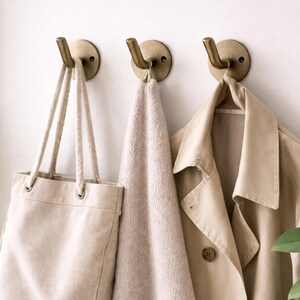 Peut inclure: Trois crochets muraux en laiton retiennent un sac fourre-tout beige, une serviette beige clair et un trench-coat beige. Les crochets sont montés sur un mur blanc, avec des plantes vertes au premier plan.