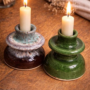 Peut inclure: Deux bougeoirs en céramique avec des bougies allumées. L'un est marron et sarcelle, l'autre est vert. Les bougies sont blanches et cylindriques. Les bougeoirs sont sur une surface en bois.