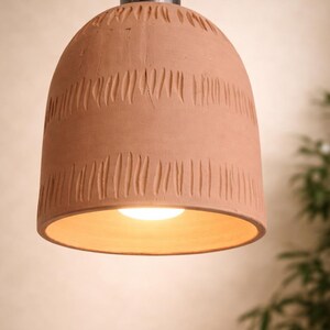 May include: A terracotta pendant light with a carved, textured pattern. The bell-shaped light has a metal fitting and a black cord. Warm light emanates from the base, illuminating the interior. The light is suspended from a black cord.