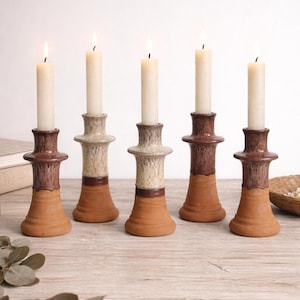 May include: Five ceramic candle holders with lit white candles. The candle holders have a terracotta base, a brown middle section, and a cream-colored top. The candles are tall and slender, with a soft glow.