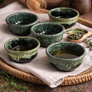 Peut inclure: Cinq petits bols en céramique faits à la main avec un glaçage vert foncé, chacun rempli d'un liquide. Les bols sont posés sur un plateau tressé avec un tissu en lin, à côté d'une cuillère en bois avec des feuilles de thé et un fouet en bambou.