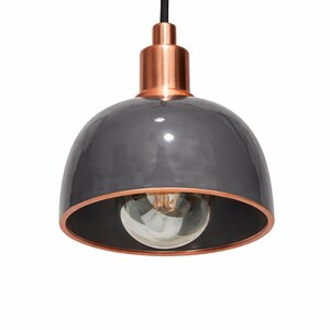 Pode incluir: Uma moderna luminária pendente com uma cúpula cinza brilhante e detalhes em cobre. A luminária tem um cabo preto e uma lâmpada visível no estilo Edison. O interior da cúpula é preto.
