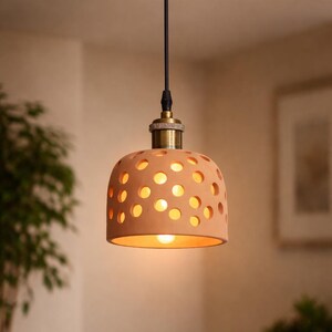 Puede incluir: Lámpara colgante de terracota con acabado de latón. La pantalla tiene cortes circulares. La lámpara cuelga de un cable negro. La cálida luz de la bombilla es visible.