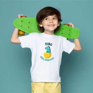 May include: A white t-shirt with a green dinosaur in an egg graphic and the text "Dino Errel Prayoga". A young person is wearing the shirt and holding a green skateboard behind their back.