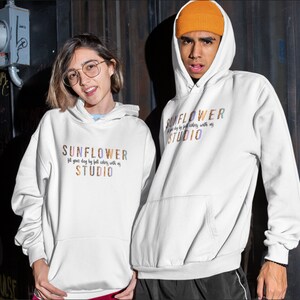 May include: Two people wearing white hoodies with the text "SUNFLOWER STUDIO" in colorful letters. The hoodies have a front pocket and a drawstring hood. The text on the hoodie reads "fill your day by full colors with us".