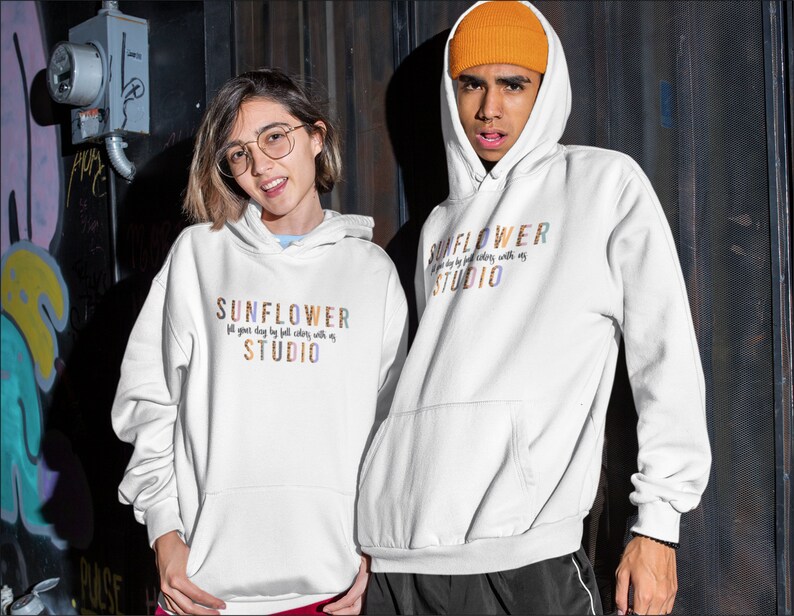 May include: Two people wearing white hoodies with the text "SUNFLOWER STUDIO" in colorful letters. The hoodies have a front pocket and a drawstring hood. The text on the hoodie reads "fill your day by full colors with us".
