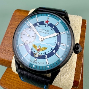 May include: A black wristwatch with a light blue face featuring the text "МОРСКАЯ АВИАЦИЯ" and a red star. The watch has a black leather band and a small seconds dial. The watch is displayed on a wooden stand.