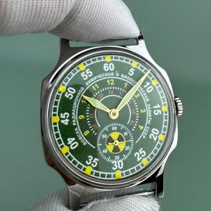 Puede incluir: Un reloj de pulsera de acero inoxidable con esfera verde y n&uacute;meros amarillos. La esfera del reloj presenta m&uacute;ltiples esferas y texto en escritura cir&iacute;lica. El reloj tiene manecillas doradas y un s&iacute;mbolo de radiaci&oacute;n en la parte inferior. El reloj lo sostiene una persona con un guante blanco.