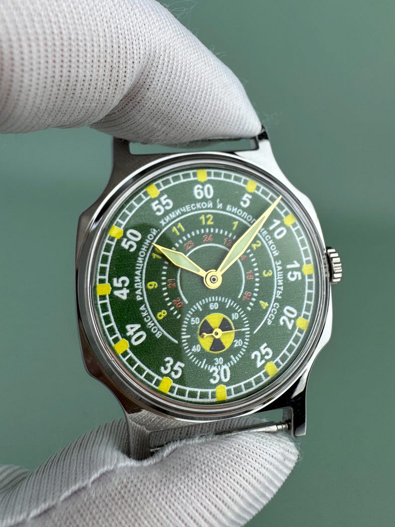 Puede incluir: Un reloj de pulsera de acero inoxidable con esfera verde y n&uacute;meros amarillos. La esfera del reloj presenta m&uacute;ltiples esferas y texto en escritura cir&iacute;lica. El reloj tiene manecillas doradas y un s&iacute;mbolo de radiaci&oacute;n en la parte inferior. El reloj lo sostiene una persona con un guante blanco.