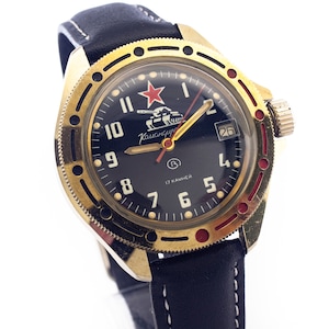 Puede incluir: Un reloj de pulsera dorado con una esfera negra y una estrella roja. El reloj tiene una correa de cuero negra y una ventana de fecha en la posición de las 3 en punto. La esfera del reloj presenta un tanque y el texto "Kauscungun" en cirílico.