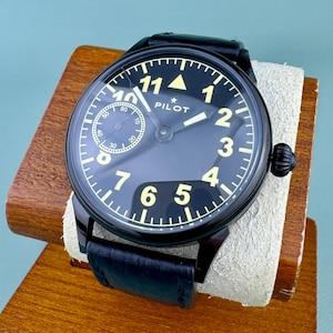 May include: Black pilot watch with a black leather band. The watch face is black with white numerals and hands. The watch is displayed on a wooden stand with a cream-colored cushion.