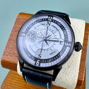 May include: A black wristwatch with a compass design on the face, featuring the text "АРКТИКА" and "СЕВЕРНЫЙ ПОЛЮС". The watch has a black leather band and is displayed on a wooden stand.