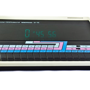 May include: A black and white digital clock with a green display showing the time 0:45:56. The clock has a label that reads "ЧАСЫ-ПРOΓΡΑΜΜΑΤΟР ЭЛЕКТРОНКА 21-10". The clock has a series of buttons with labels in Cyrillic.
