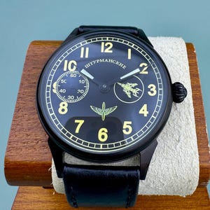 May include: A black wristwatch with a black leather band. The watch face is black with gold numerals and hands. The watch has the word "ШТУРМАНСКИЕ" and a small airplane graphic on the face. The watch is displayed on a wooden stand.