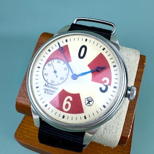 May include: Vintage wristwatch with a cream-colored face featuring red and white accents, black numerals, and blue hands. The watch has a black fabric band and is displayed on a wooden stand. The watch face includes the text "Made in USSR".